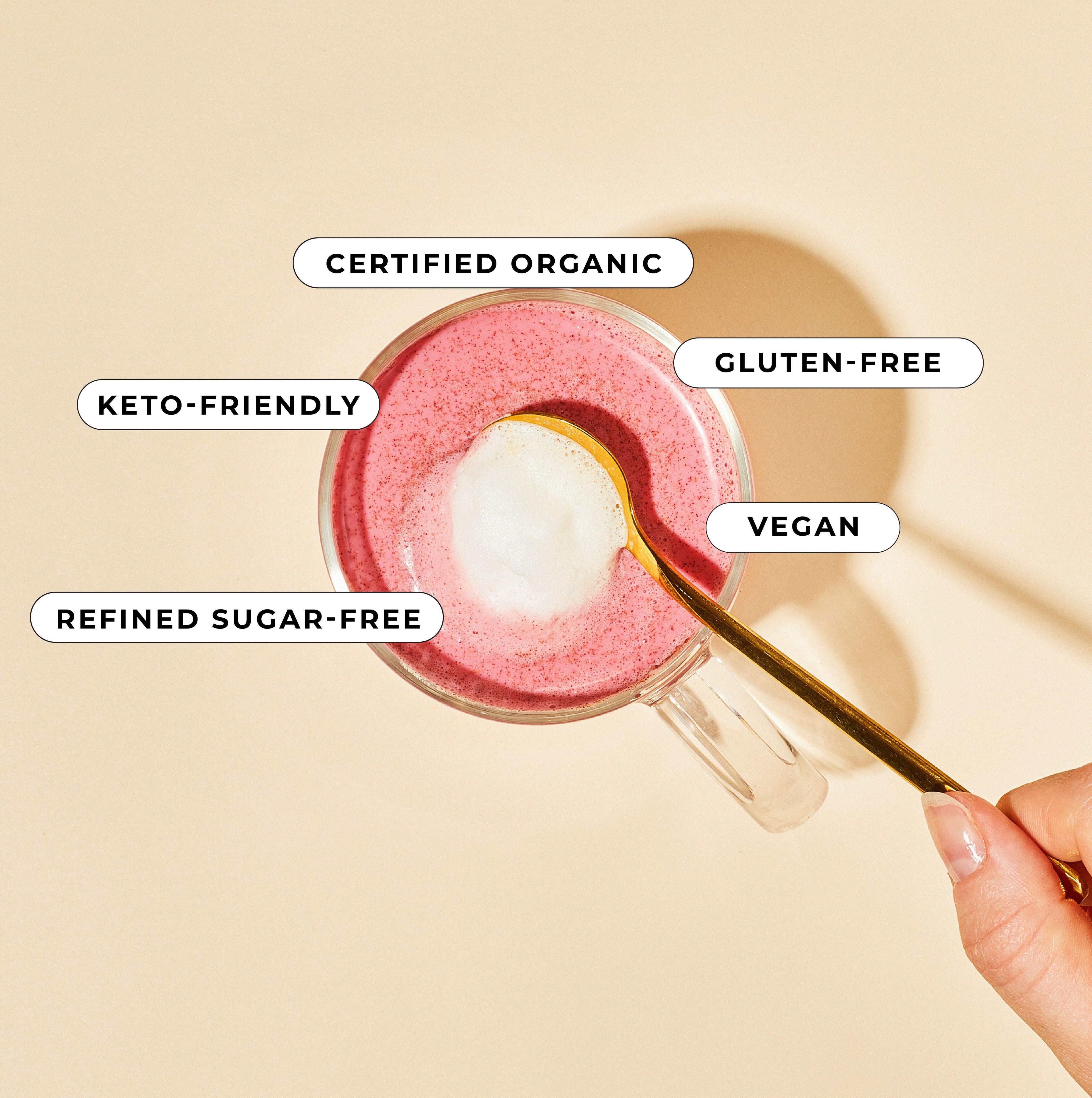 A hand stirs a pink Beetroot Blend latte by Blume in a clear mug, topped with foam. Its labeled as certified organic, gluten-free, vegan, keto-friendly, and refined sugar-free—a perfect caffeine-free boost made with organic ingredients.