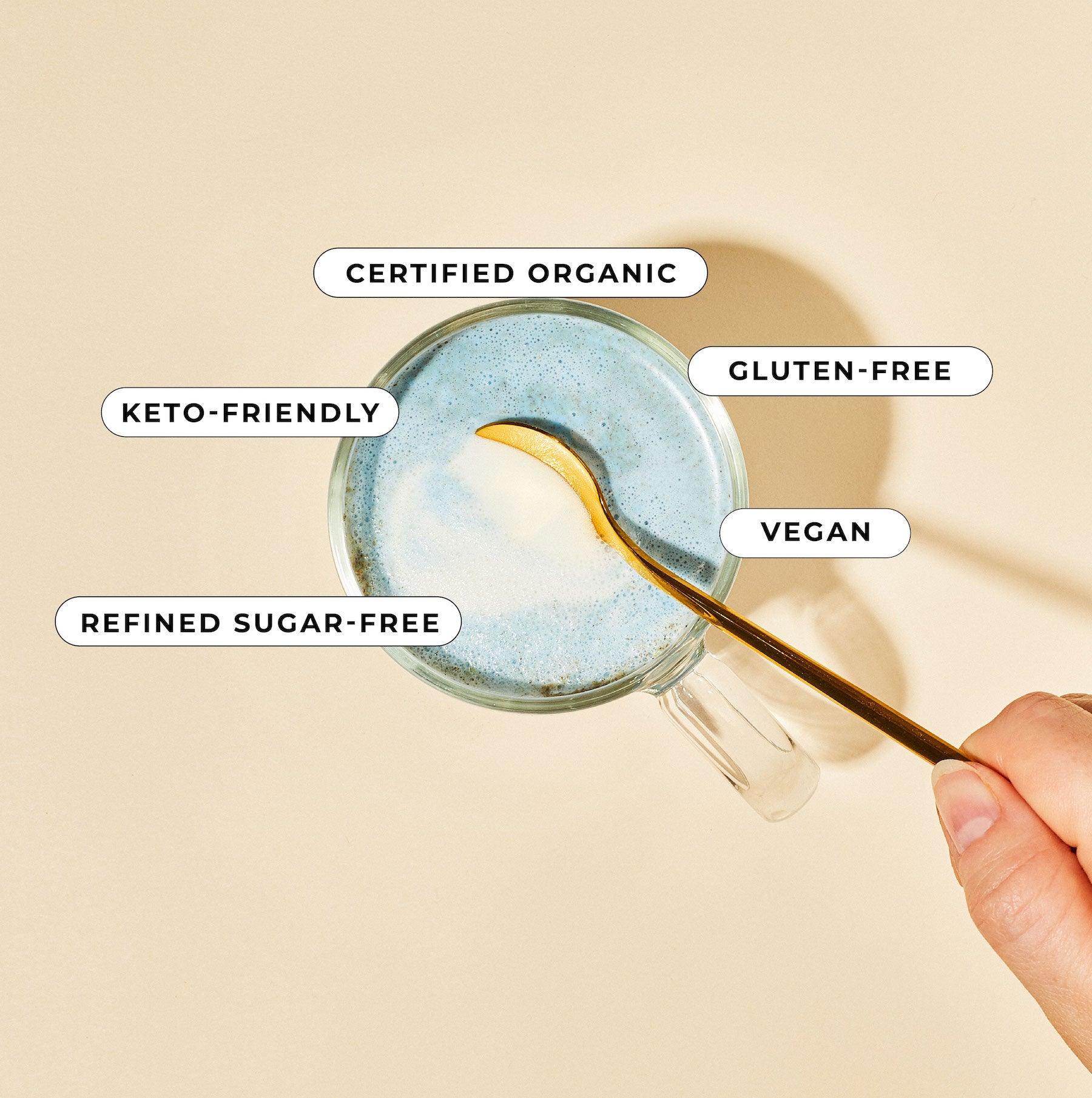 A person holds a spoon over a glass with Blumes Blue Lavender Blend, showcasing its soothing blue hue. Labels read: Certified Organic, Keto-friendly, Refined Sugar-free, Gluten-free, and Vegan. A light beige background enhances its calming allure.