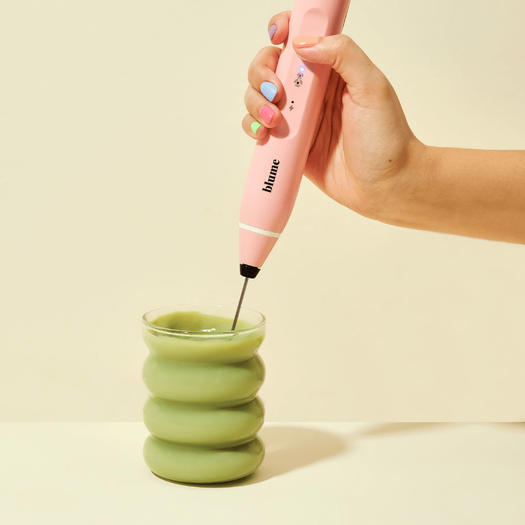 A hand with pastel-painted nails holds a pink handheld milk frother above a bubble glass filled with green liquid, likely an iced latte. The wavy-shaped Blume Bubble Cup stands out against the neutral beige background, adding a whimsical touch.