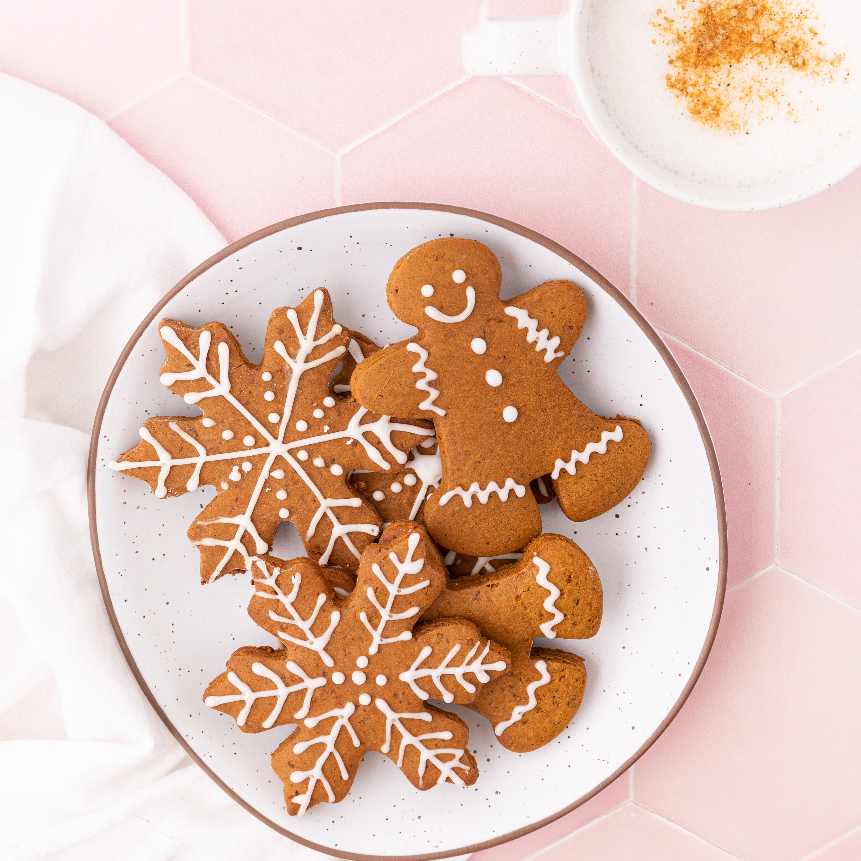 A plate with gingerbread cookies, featuring a cheerful gingerbread person and snowflakes with white icing, rests on a pink hex tile. Nearby, a cozy cup of mint cocoa enhances the mood beside a white cloth. Discover similar treats in Our Holiday Recipe E-Book by Blume.