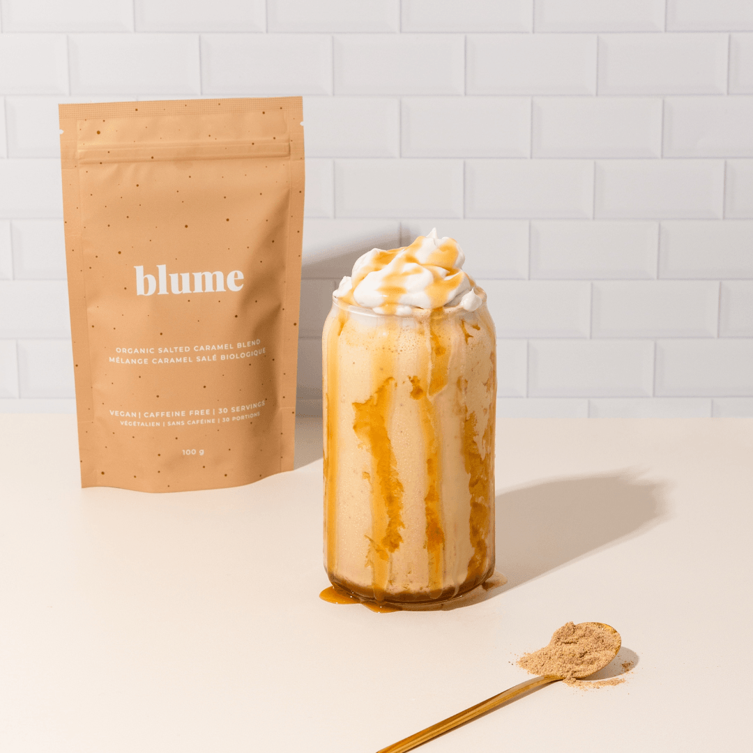 A glass of creamy caramel latte with whipped cream and caramel drizzle sits on a white tiled surface. Behind it, a peach-colored Blume bag labeled Salted Caramel Blend highlights its superfood ingredients. A gold spoon with the latte powder is placed nearby.