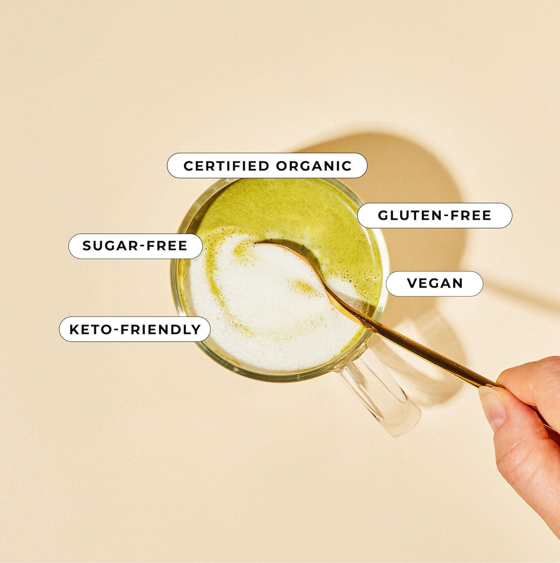 A hand stirs a vibrant green Matcha Coconut Blend by Blume in a transparent mug with a gold spoon against a beige background. Labels around the mug proudly display Certified Organic, Gluten-Free, Vegan, Sugar-Free, and Keto-Friendly.