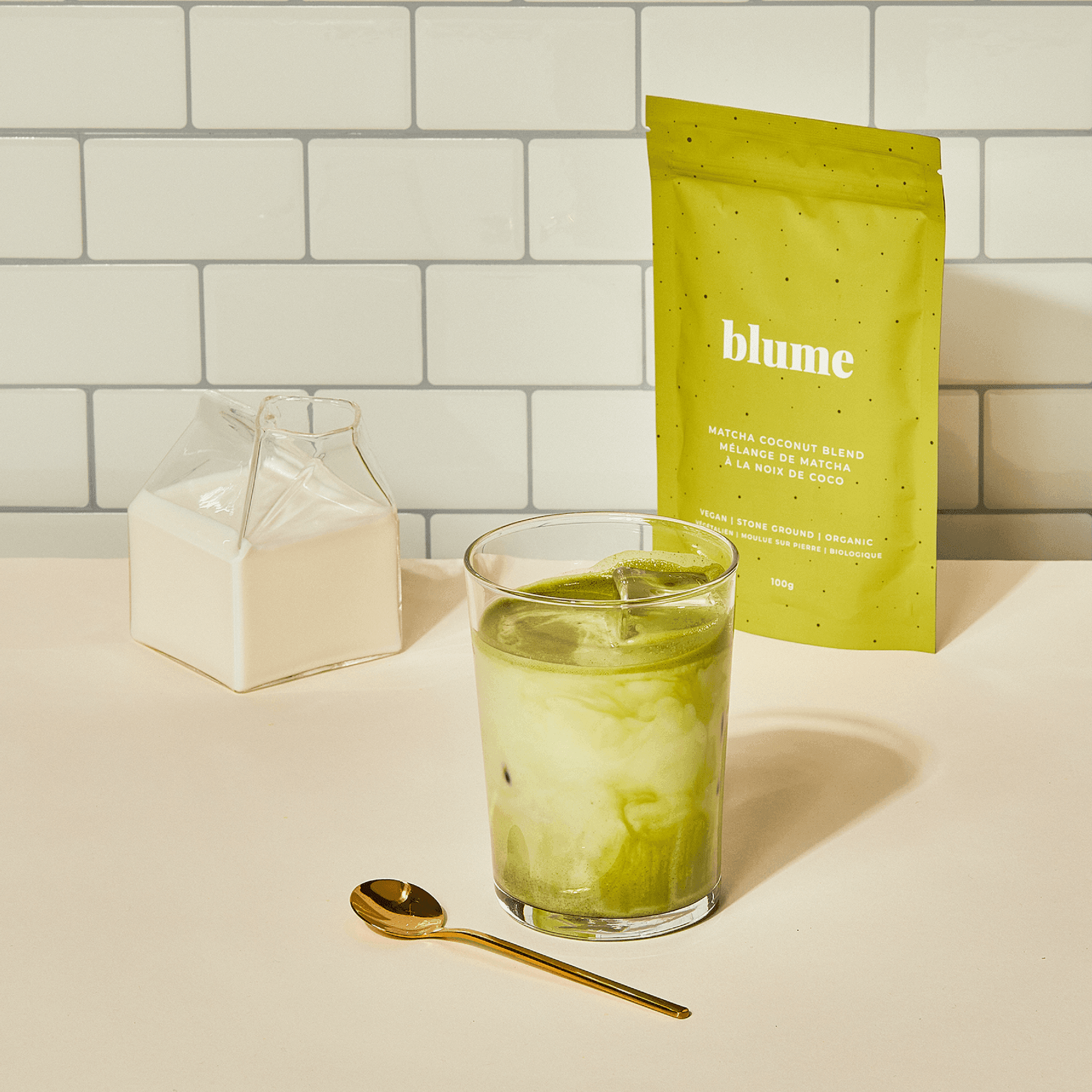 On a light surface, a glass of iced matcha latte with antioxidant-rich moringa rests beside a small golden spoon, a milk carton, and Blumes green Matcha Coconut Blend package. The background has white tiled walls.