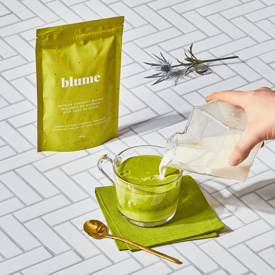 A hand pours milk from a glass pitcher into a cup of green Matcha Coconut Blend by Blume, known for its antioxidants, on a green napkin. Beside the cup lies a gold spoon. In the background, theres a green Blume package and a small vase with spiky plants on the tiled surface.