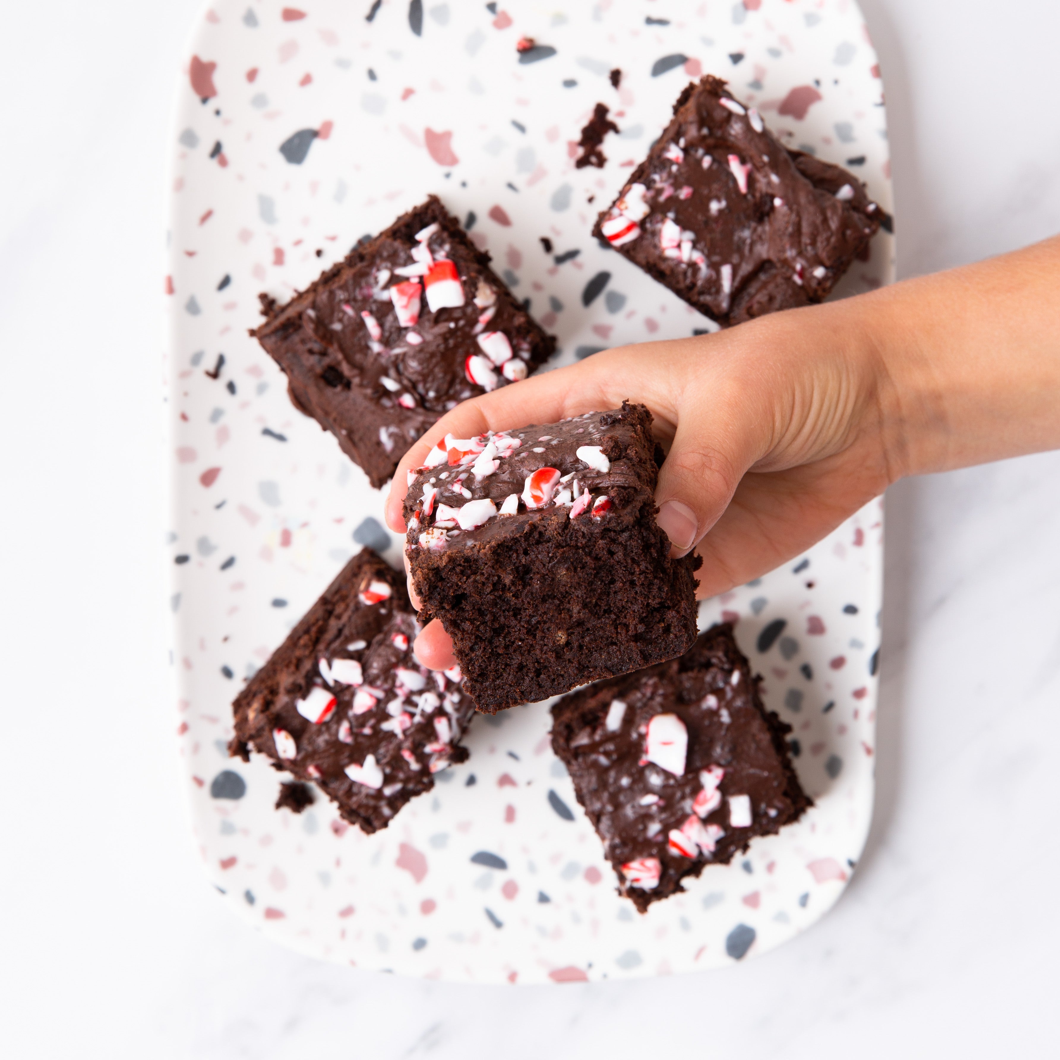 A hand holds a chocolate brownie infused with mint cocoa and topped with crushed candy canes above a speckled plate of four similar treats. The scene sits on a white marble surface, evoking the festive spirit found in Blumes Our Holiday Recipe E-Book.