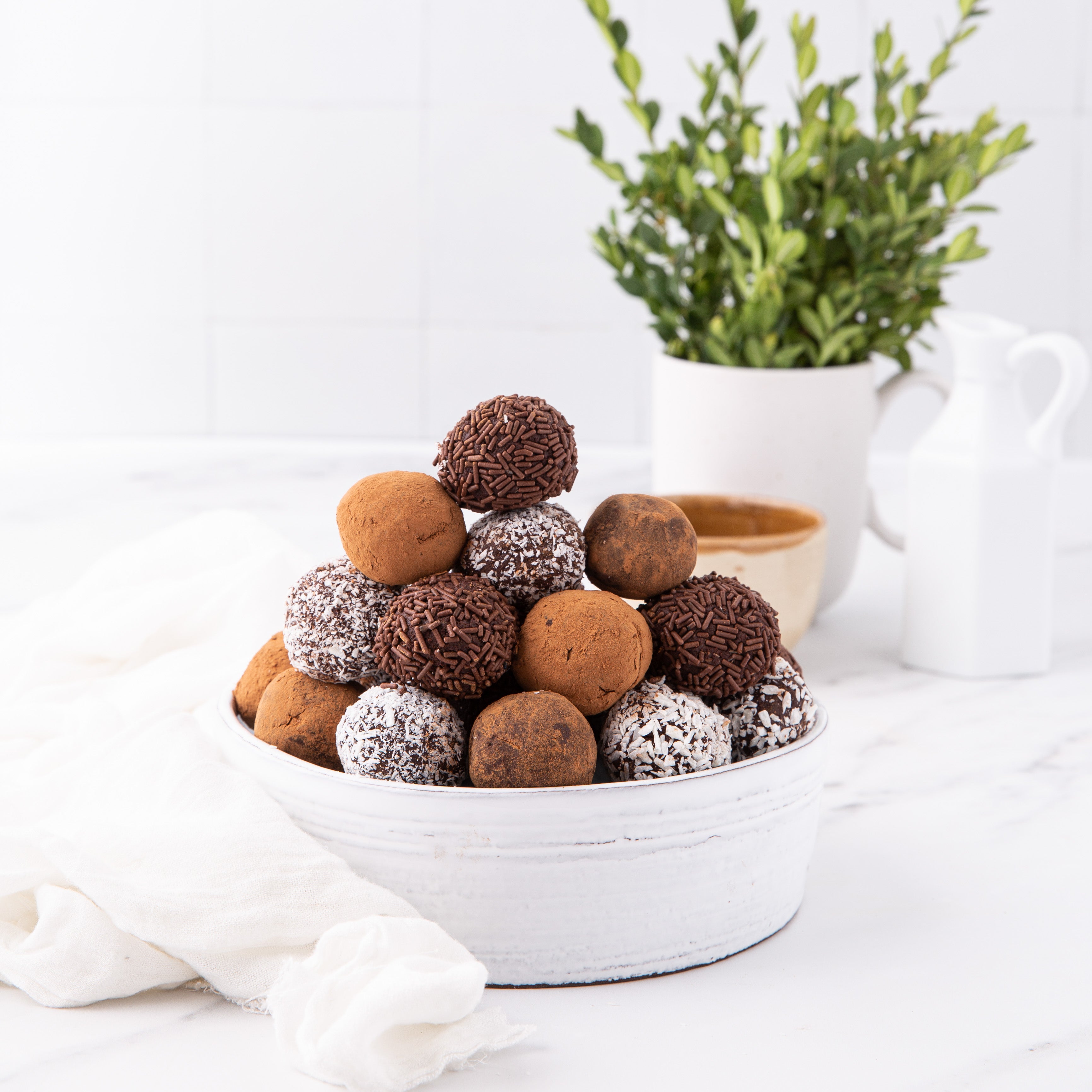 A white bowl of assorted chocolate truffles with cocoa, sprinkles, and coconut flakes sits on marble. In the background is a white vase with green leaves and a mint cocoa cup, setting a cozy scene that echoes the delights in Blumes Our Holiday Recipe E-Book.