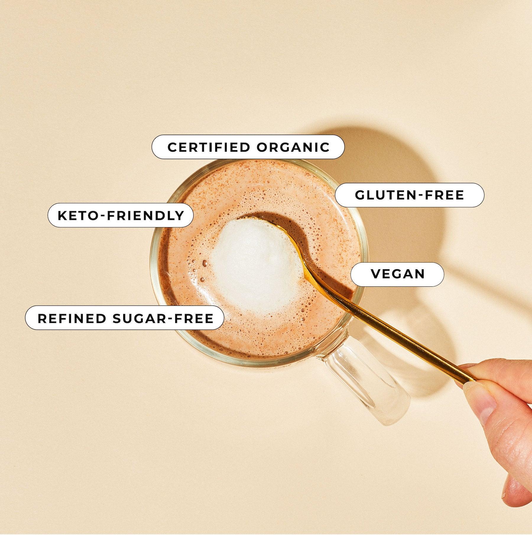 A hand stirs a frothy Reishi Hot Cacao Blend by Blume in a clear mug labeled Certified Organic, Keto-Friendly, Refined Sugar-Free, Gluten-Free, and Vegan. A light beige background adds warmth to this adaptogen-rich moment.