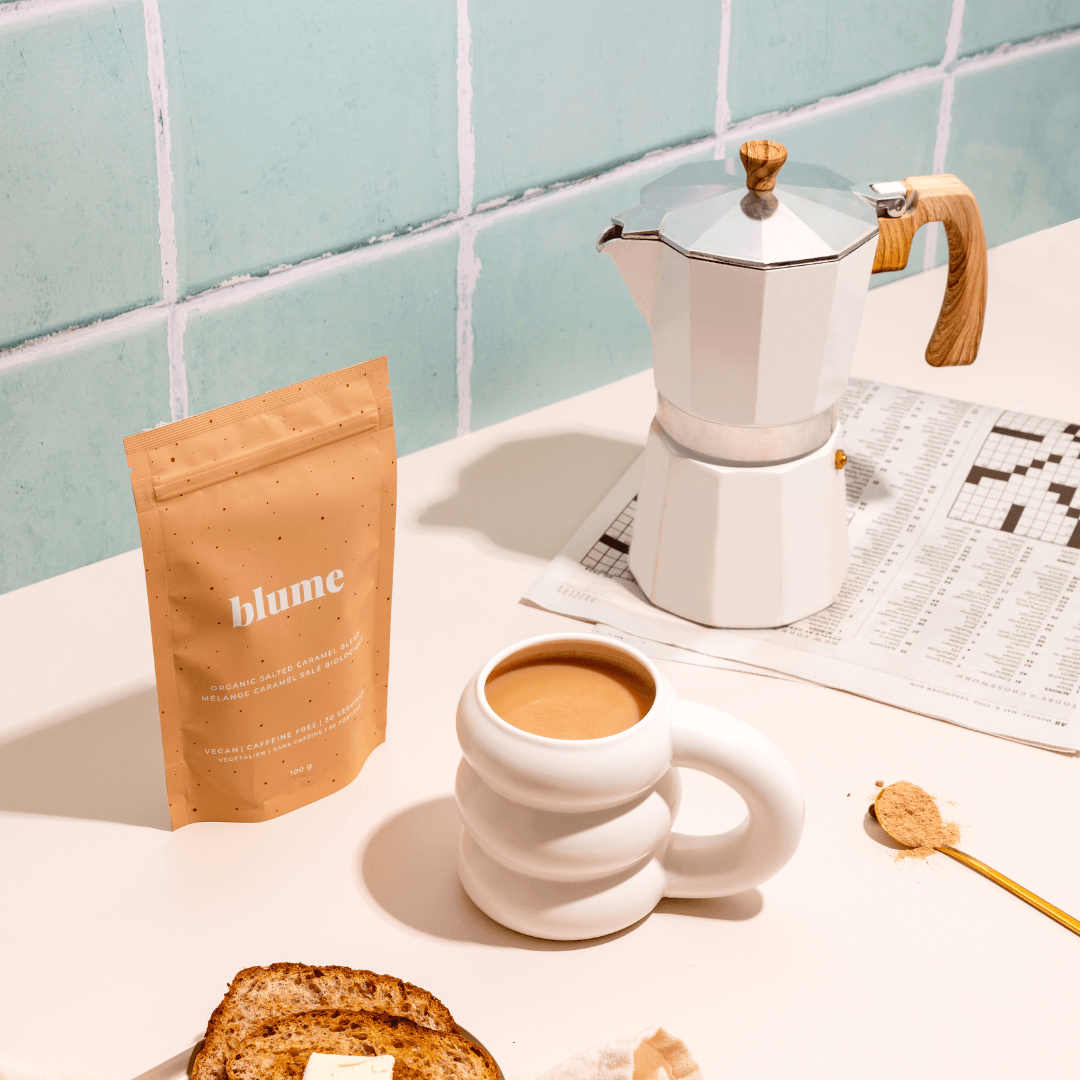 A kitchen scene features a kettle, a crossword puzzle, and a cup of coffee next to a packet labeled Blume Salted Caramel Blend. Nearby, toast with spread and a spoon with coffee powder rest on a light surface framed by tiled backsplash.