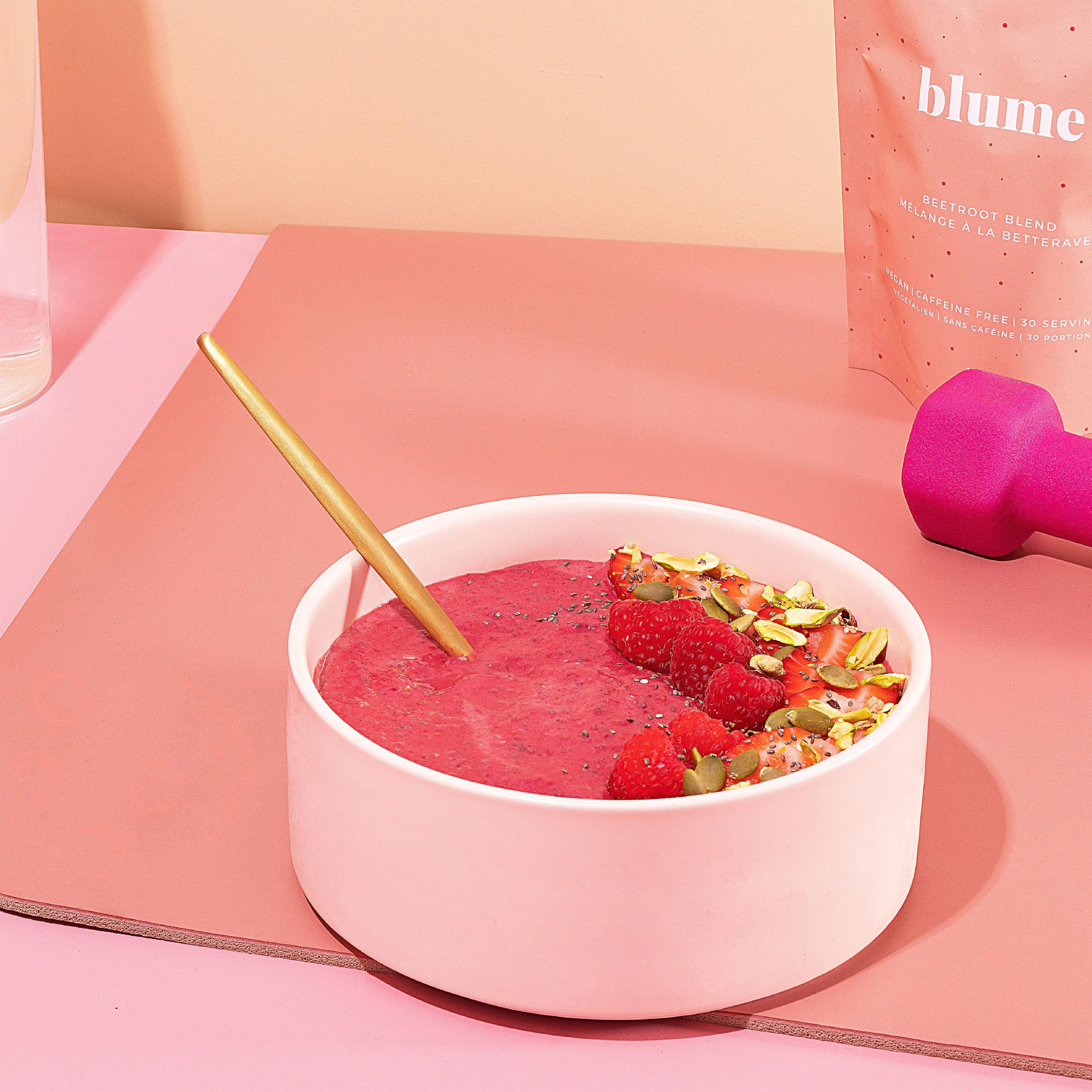 A vibrant pink smoothie bowl featuring Blumes organic Beetroot Blend is topped with raspberries, pistachios, and sliced strawberries. A gold spoon rests amidst a pink backdrop with a dumbbell and part of the product package. Ideal for a caffeine-free boost!.
