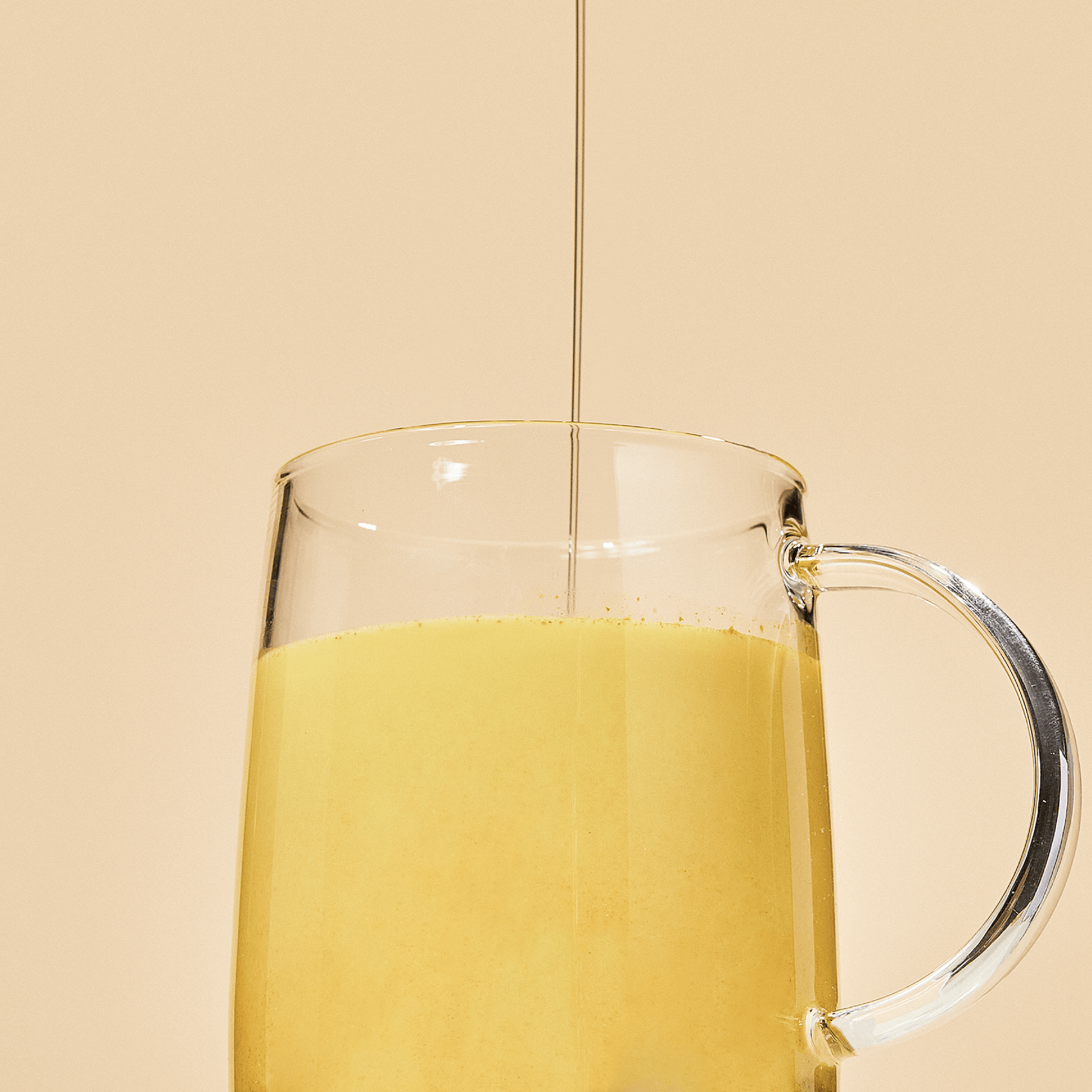 A glass mug filled with Blumes Turmeric Blend, a creamy yellow latte, is set against a beige background. A thin stream of liquid pours into the center of the drink, casting faint shadows inside.