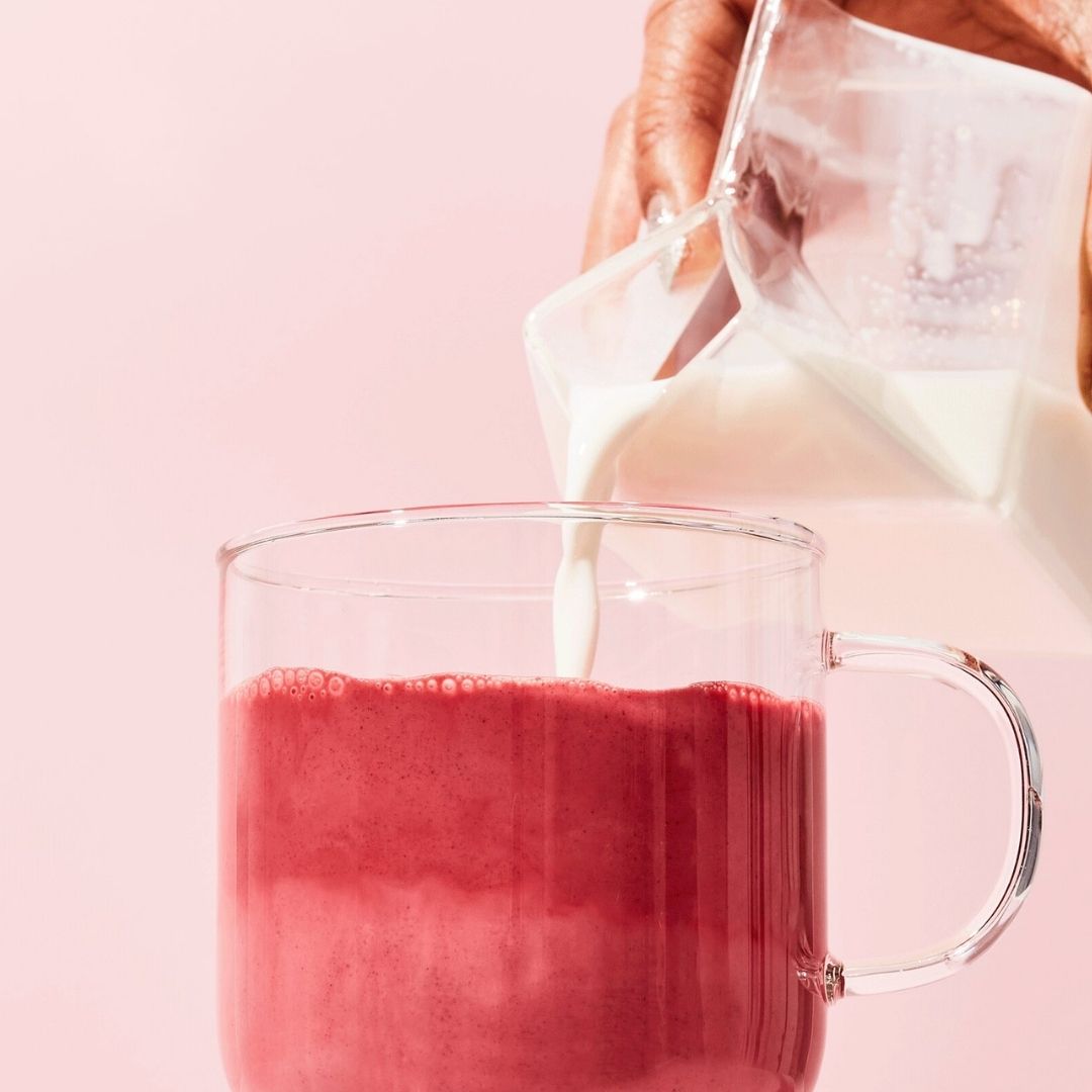 Milk pouring into a glass mug of pink latte made with Blume Beetroot Blend on a pink background