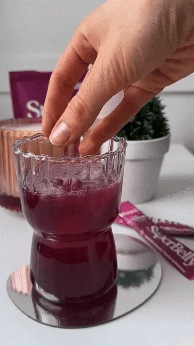 A hand stirring a glass of purple liquid with ice cubes. The glass is placed on a small reflective surface. In the background, there are a plant and purple packaging.