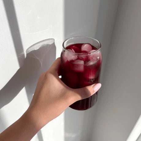 A hand holding a glass of iced red drink against a light background, with soft shadows cast by sunlight.