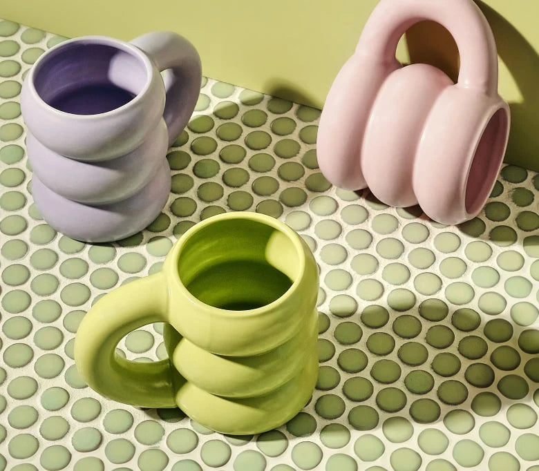 Three ceramic mugs with a twisted, spiral design sit on a patterned surface. The mugs are in pastel pink, green, and lavender colors, complementing the light green background.