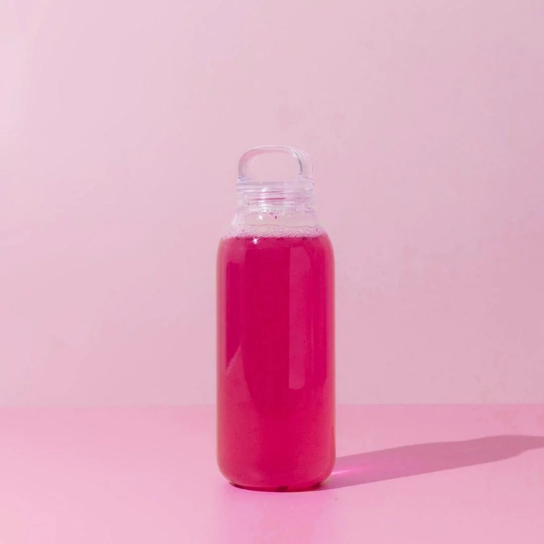 A clear bottle filled with pink liquid stands on a light pink surface against a matching background. The bottle has a simple, rounded top, and the liquid reaches near the top. A shadow is cast on the surface to the right.