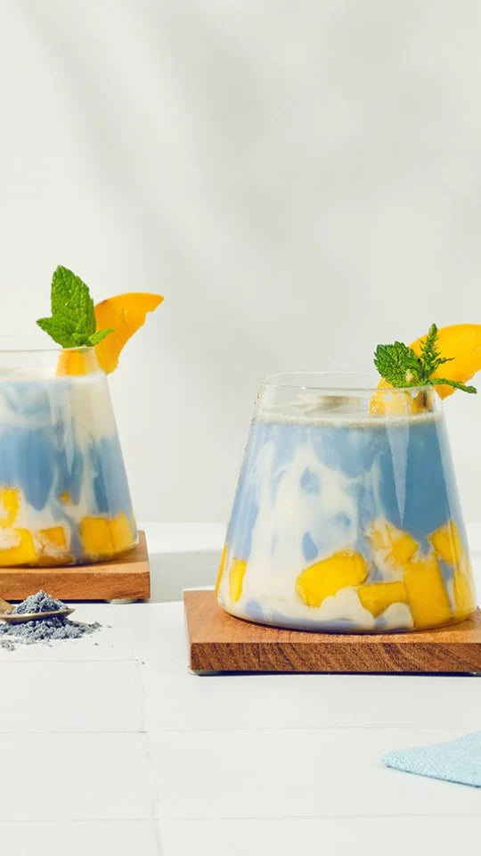 Two glasses filled with a marbled blue and white creamy drink, garnished with mint leaves and orange slices. The glasses have layers of yellow fruit chunks and sit on wooden coasters. A small pile of blue powder is visible nearby on a white surface.