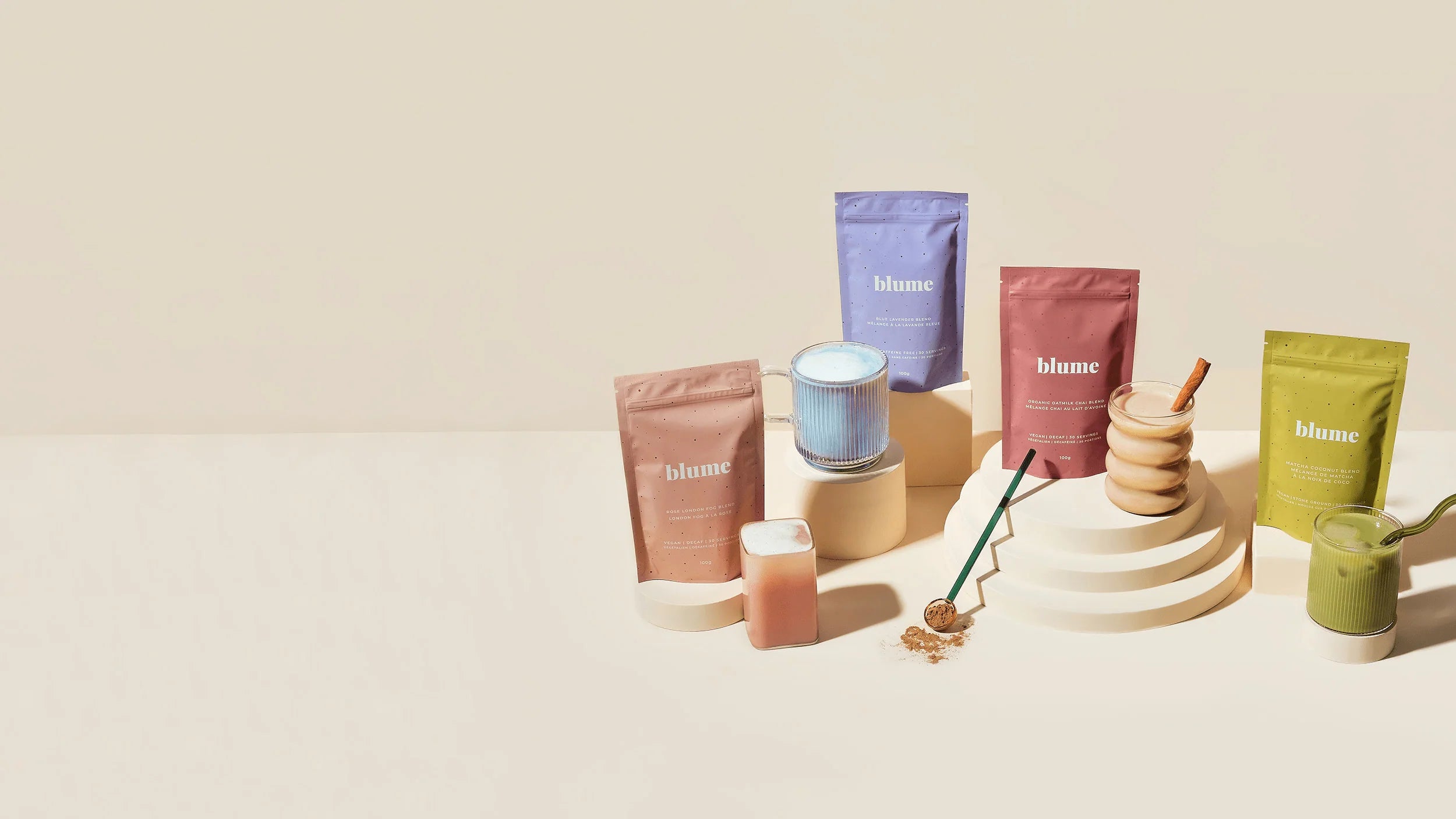 A display of Blume products on beige pedestals, including colorful packets, a blue latte in a glass, and a green smoothie. Some powders are spilled with a spoon next to them. The background is a light cream color.