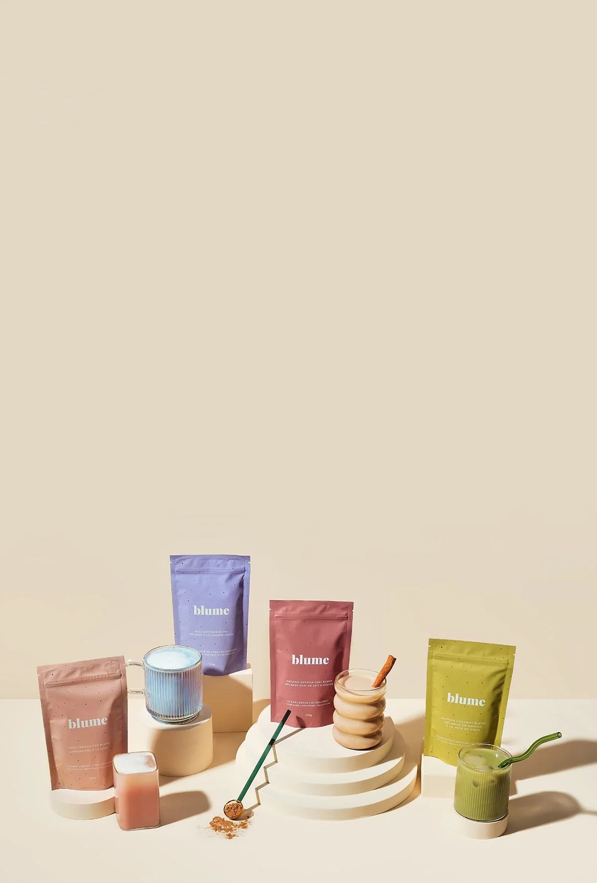 Various colorful Blume products including powder pouches, ceramic jars, and a honey dipper arranged on a beige background with minimalist cylindrical props.