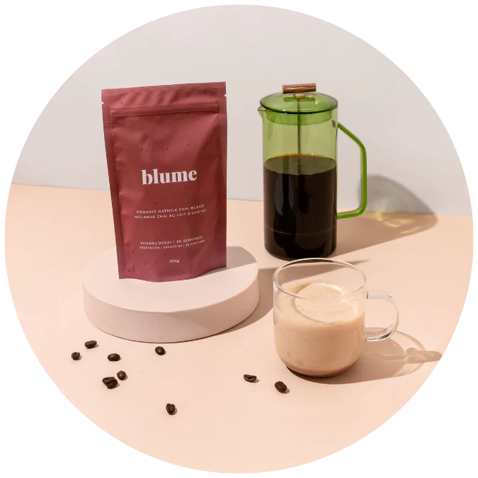 A pink packet of Blume organic coffee is displayed on a white pedestal. Nearby, a glass French press contains brewed coffee, and a glass cup holds creamy coffee. Coffee beans are scattered on the light surface.