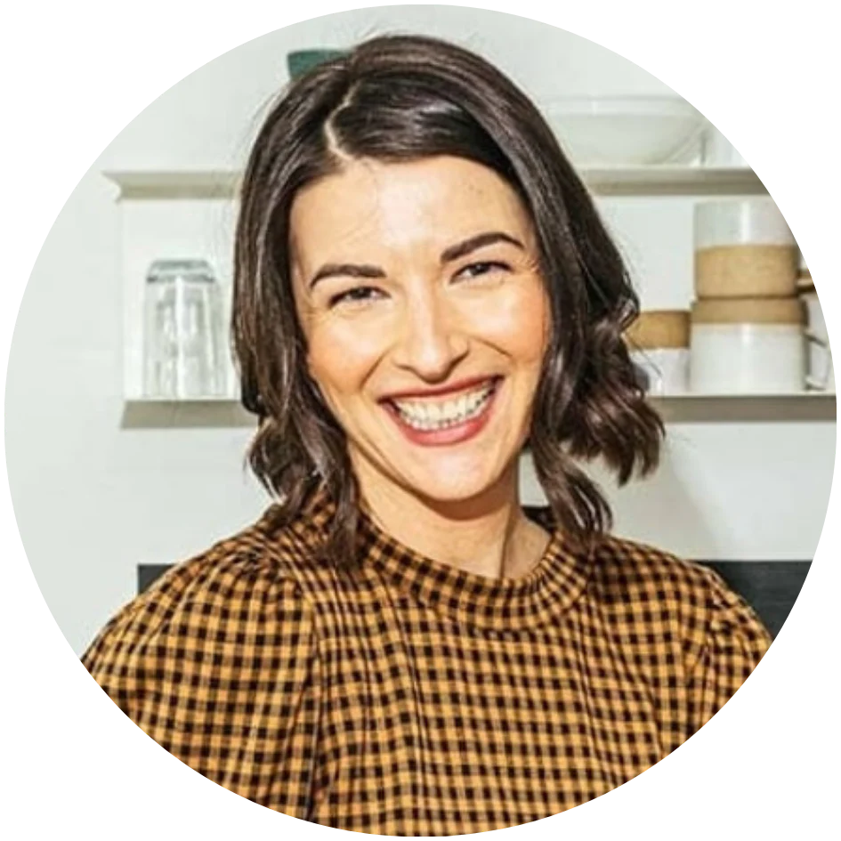 Smiling person with short brown hair wearing a brown checked shirt. They are in an indoor setting with kitchen shelves in the background.