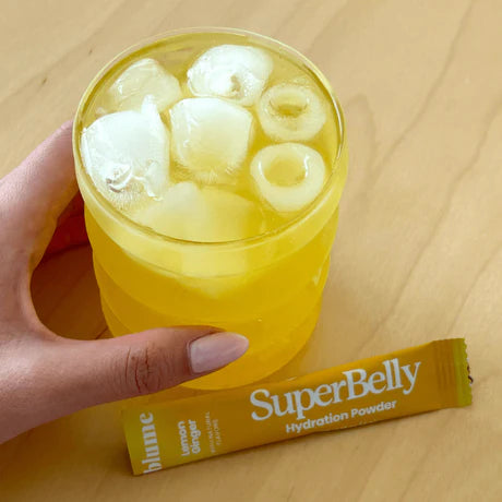 A hand holding a yellow drink with ice cubes in a textured glass on a wooden table. Next to the glass is a sachet labeled SuperBelly Hydration Powder in lemon ginger flavor.