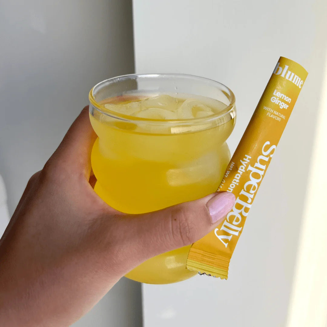 A hand holding a rounded glass filled with yellow liquid and ice cubes. Next to the glass, theres a yellow packet labeled Blume Supervitaly Hydration in lemon ginger flavor.