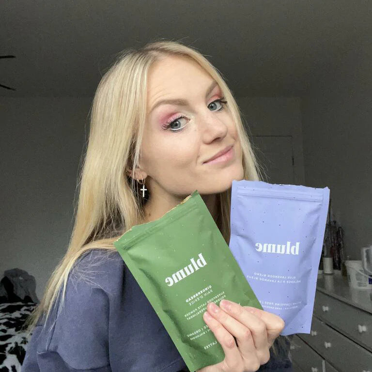 A person with long blonde hair is smiling and holding two packages labeled Blume. One package is green, and the other is light purple. The person is wearing a black top and standing in a room with white walls and a dresser.