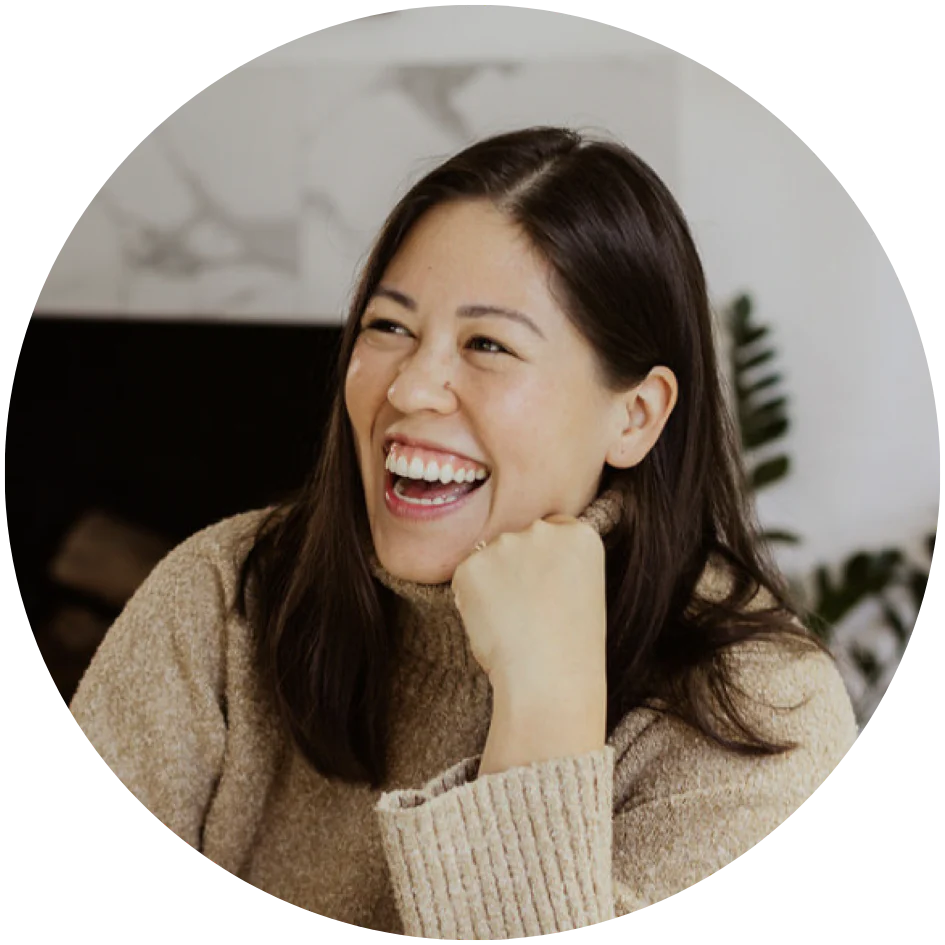 A woman with long dark hair and a beige sweater smiles joyfully. She rests her chin on her hand in a relaxed manner. A blurred background with abstract shapes and green foliage is visible.