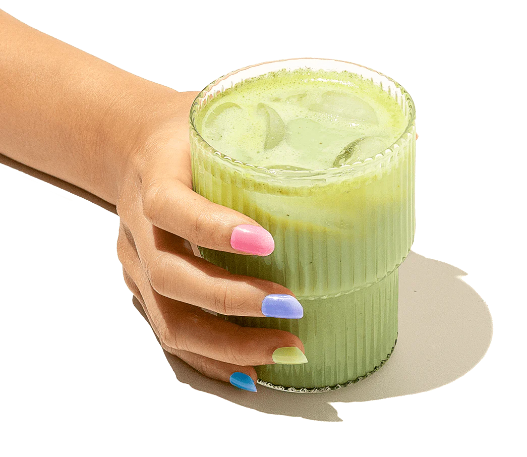 A hand with multicolored fingernails holds a ribbed glass filled with a green iced drink.