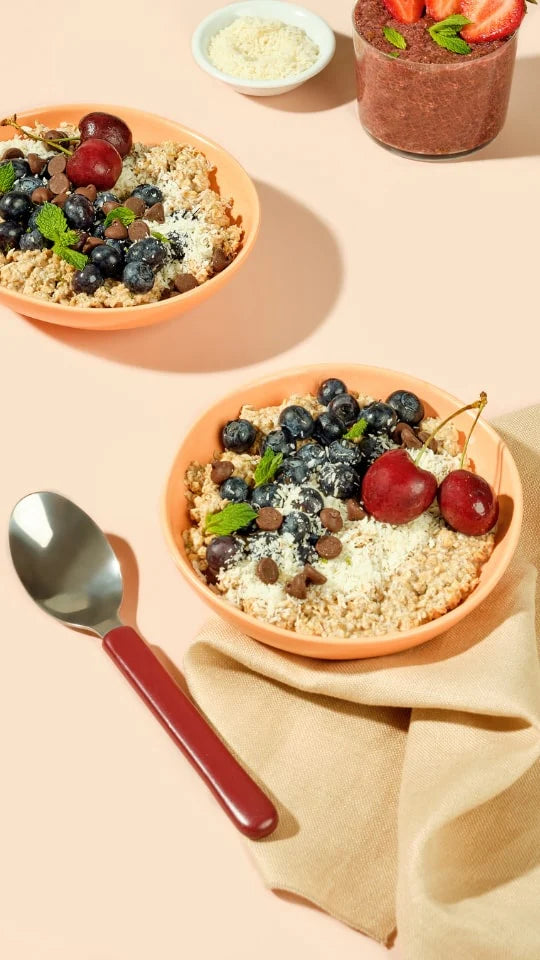 Two bowls of oatmeal topped with blueberries, cherries, mint leaves, and seeds sit on a light surface. A metal spoon with a red handle is nearby. In the background are a small dish of seeds and a smoothie topped with sliced strawberries.