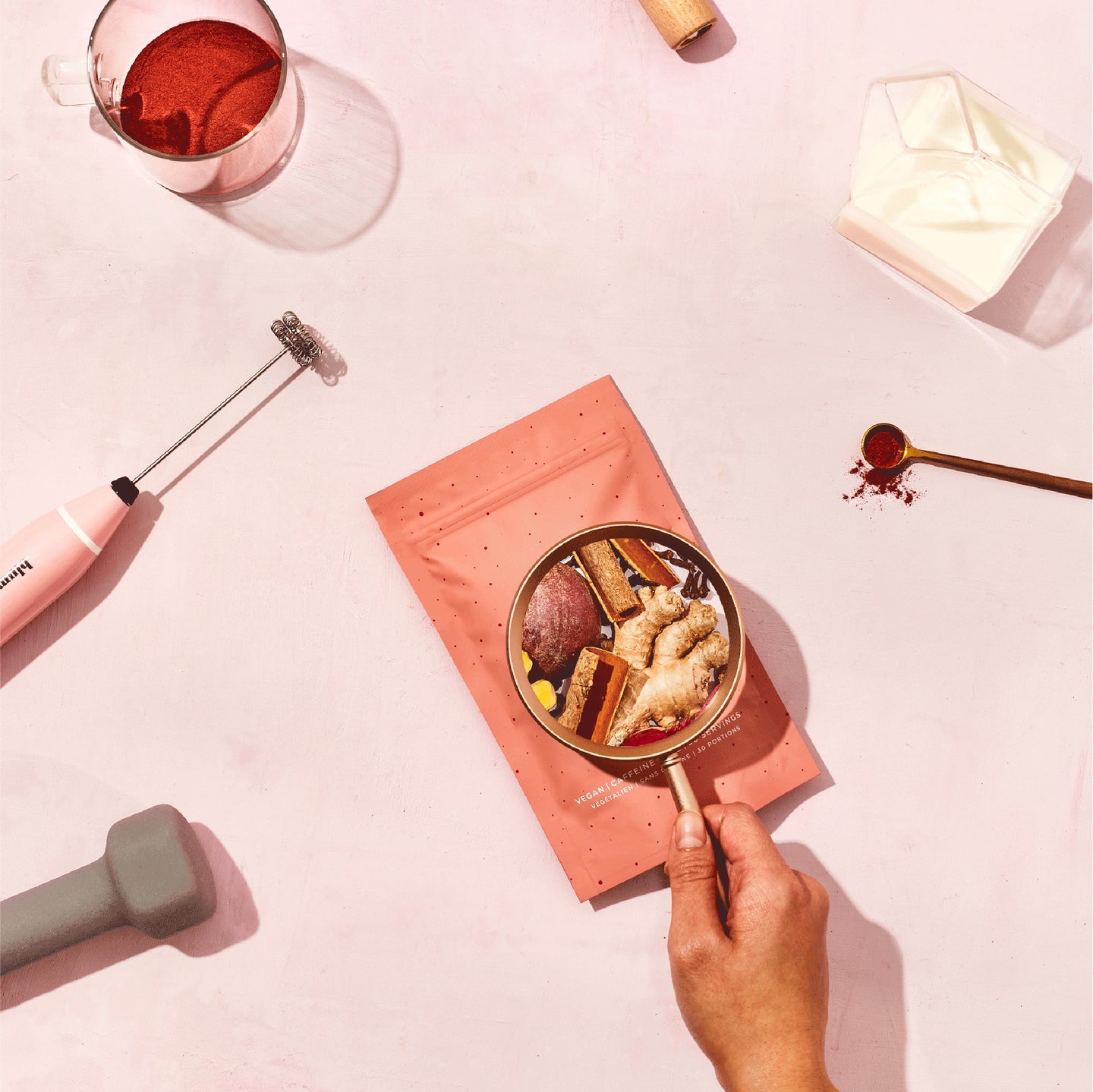 A bag of Blume Beetroot Blend surrounded by a Blume Pink Milk Frother, grinds in a glass cup, milk in a glass milk carton creamer, a gold spoon with grinds, and a dumbbells weight