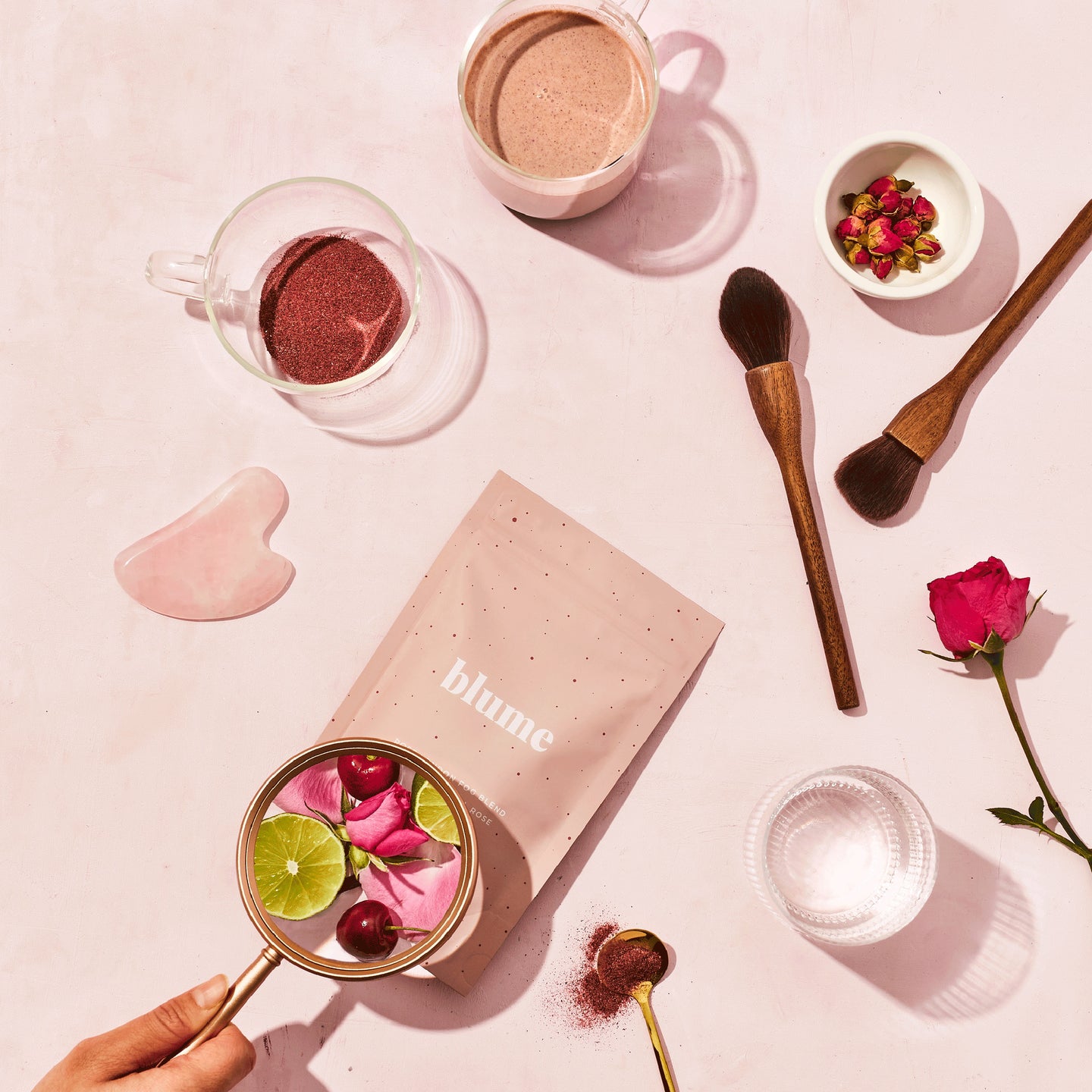 A bag of Blume Rose London Fog surrounded by glass cups, a Gua Sha, makeup brushes, flowers on a pink background 