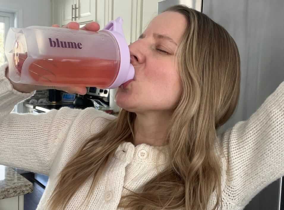 Woman drinking from a pink blume shaker cup with SuperBelly in a kitchen.