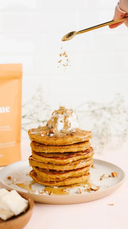 A stack of pancakes topped with whipped cream, chopped nuts, and a drizzle of syrup. A hand sprinkles more nuts from a spoon above. A package and a bowl with butter are visible in the background on a light surface.