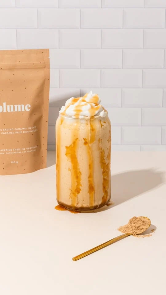 A glass filled with a creamy caramel drink topped with whipped cream and caramel drizzle. A packet labeled Blume and a small golden spoon with caramel powder are placed on a light surface against a white tiled background.