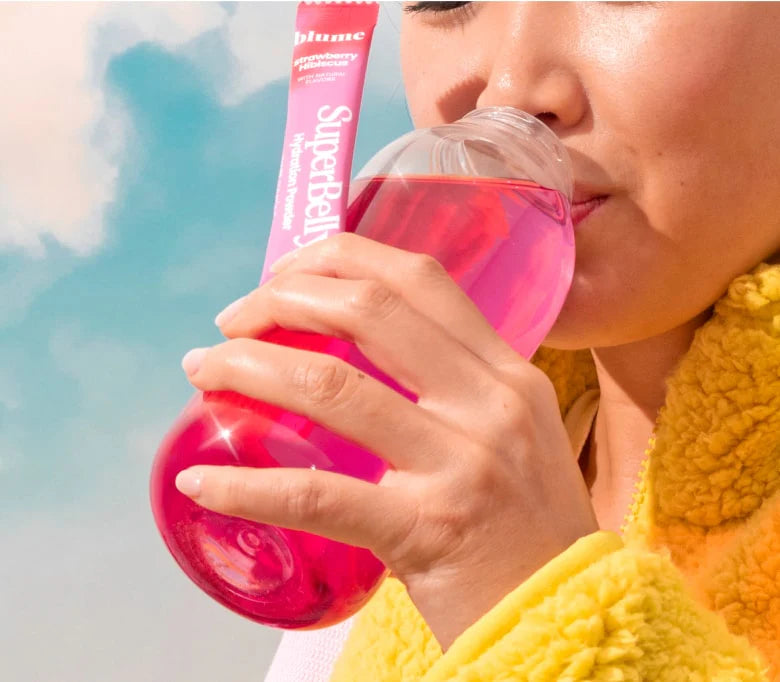 A person wearing a yellow garment sips from a clear bottle filled with pink liquid. They hold a tube labeled SuperBelly Raspberry Lemonade Flavor next to the bottle. The background is a clear blue sky with some clouds.