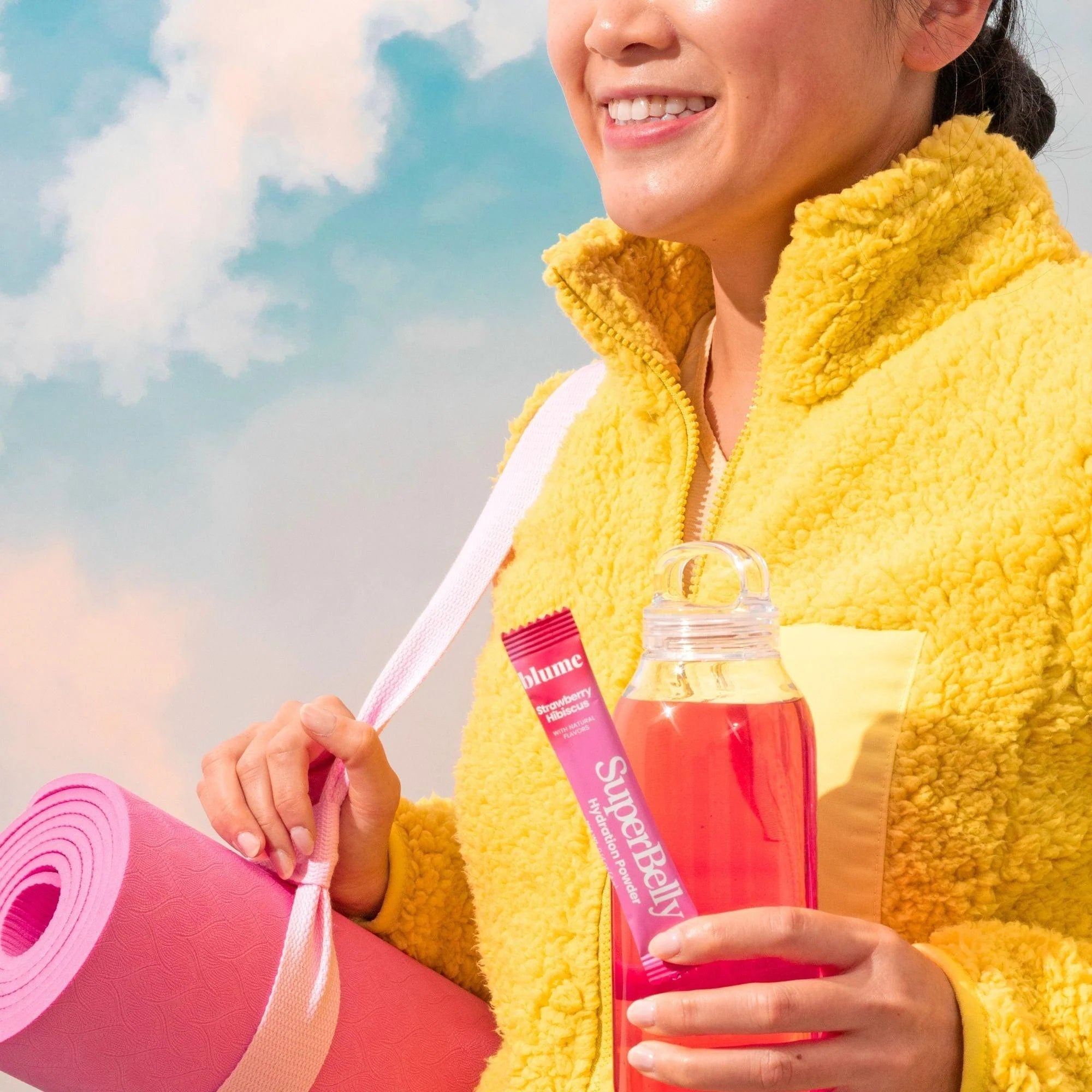 A person in a yellow fleece jacket holds a pink yoga mat and a clear bottle filled with red liquid. They are also holding a pink packet labeled SuperBelly. The sky with clouds is visible in the background.