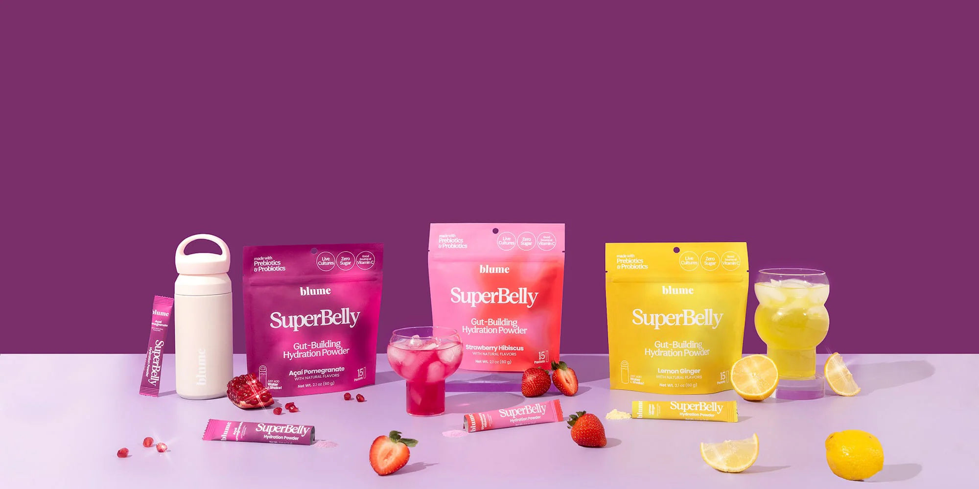 A display of SuperBelly hydration powder products on a table. Includes packets and individual sticks in red, pink, and yellow. A shaker bottle, glass with red drink, and a glass with yellow drink accompany various fruits like lemons and strawberries.