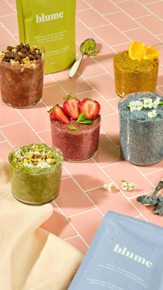 Glass jars filled with colorful smoothies topped with fruit and seeds are arranged on a pink tiled surface. A green packet labeled blume is in the background, and a blue packet is in the foreground, next to a beige cloth.