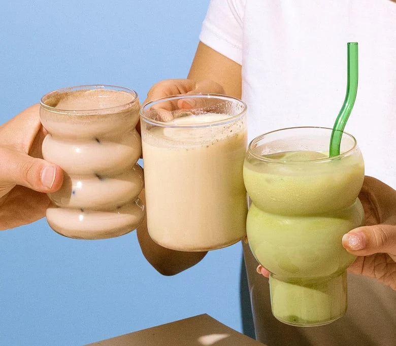 Three hands holding drinks in textured glasses against a blue background. One glass contains a chocolate beverage, another has a creamy drink, and the third holds a green drink with a green straw.