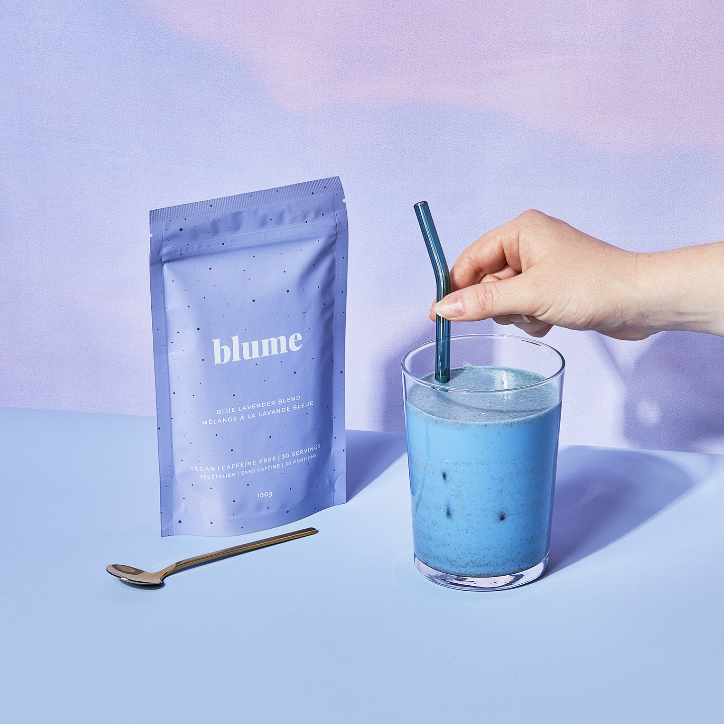 Blue Lavender latte in a glass cup, a hand putting a glass blue straw in a cup, with a bag of Blume Blue Lavender Latte and gold spoon on a purple gradient background
