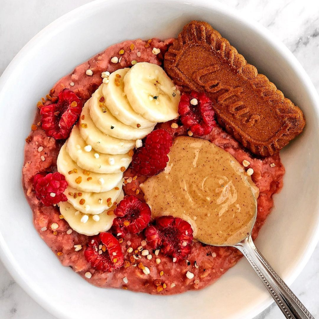 A bowl of pink overnight oats made with Blume Beetroot Blend, topped with sliced banana, raspberries, a spoon of peanut butter, and a Lotus cookie taken by @cookwithlisha