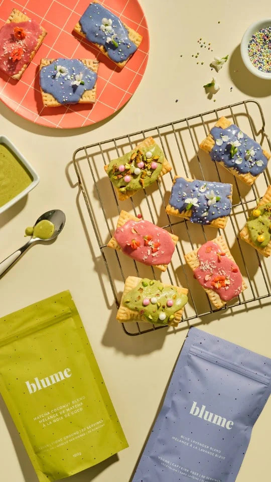 Colorful frosted pastries topped with sprinkles are displayed on plates and a wire rack. Two small bowls and a spoon with green sauce or icing are nearby. Two Blume-brand beverage mix packages in green and blue are at the bottom.