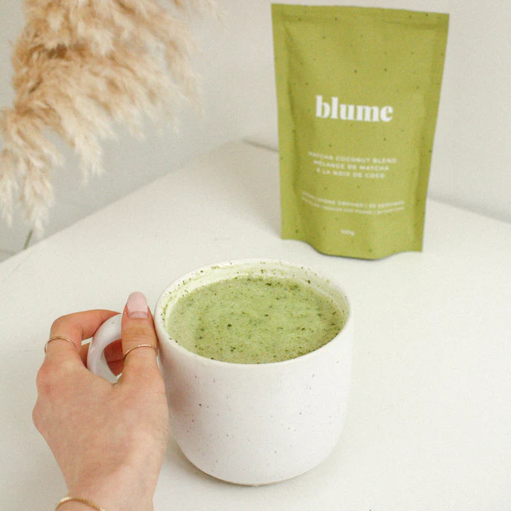 A hand holding a white mug filled with a green drink, likely a matcha latte. In the background, a green packet labeled Blume sits on a white surface, next to dried pampas grass in soft lighting.