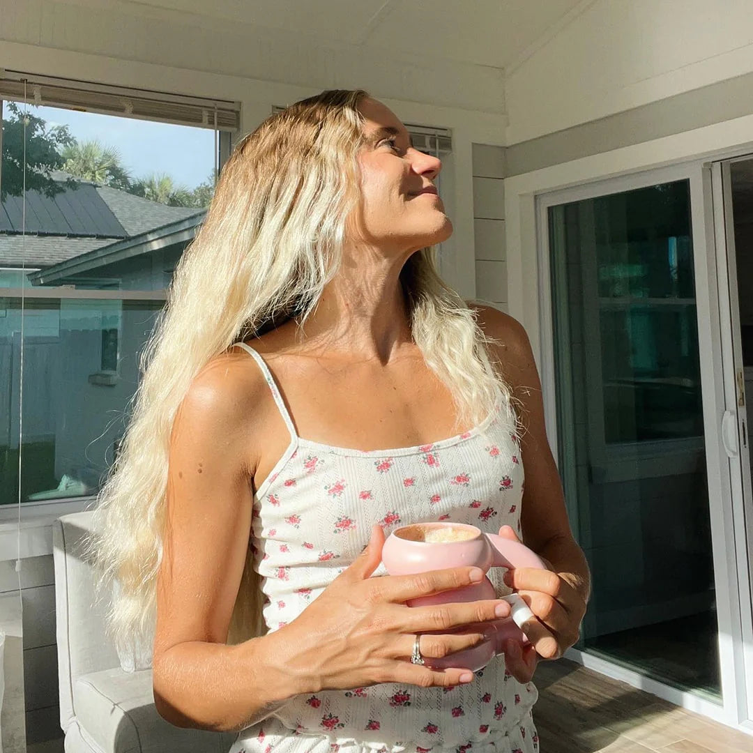 Woman with long blonde hair, wearing a white floral dress, stands in a sunlit room holding a pink mug. She is smiling and looking upwards. There are large windows and a sliding glass door behind her.