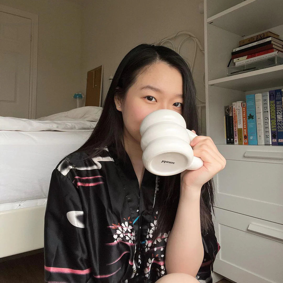 A person with long black hair is sitting on the floor near a bed, holding a large white mug with both hands. They are wearing a black robe with floral patterns. A bookshelf with various books is visible in the background.