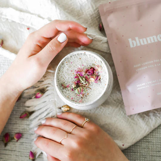 Hands with manicured nails holding a cup of frothy beverage topped with rose petals. A pink packet labeled Blume is nearby on a light-colored fabric. A gold ring is on one hand, and rose petals are scattered around.