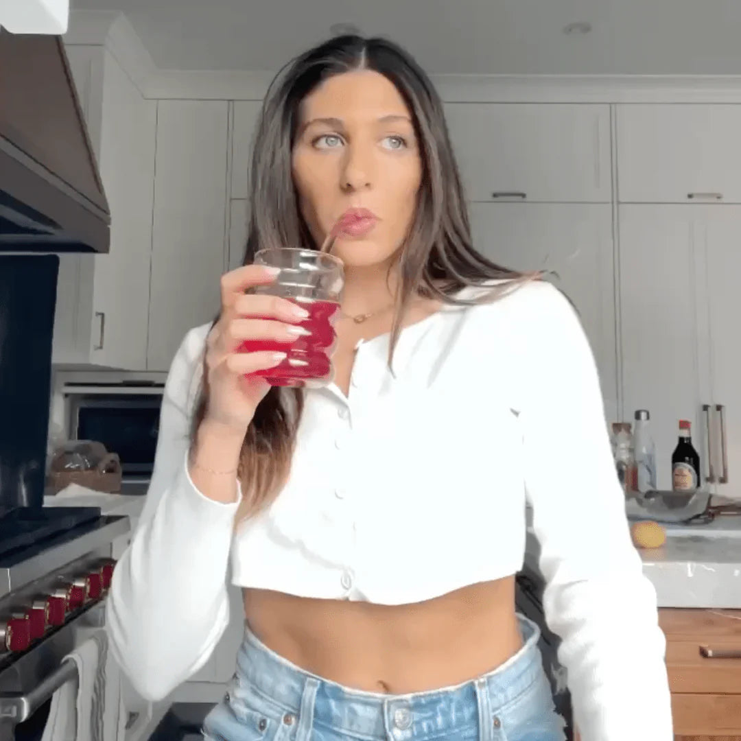 A woman with long brown hair sips a red drink through a straw in a kitchen. She is wearing a white cropped sweater and jeans, standing casually in front of the stove and kitchen cabinets.